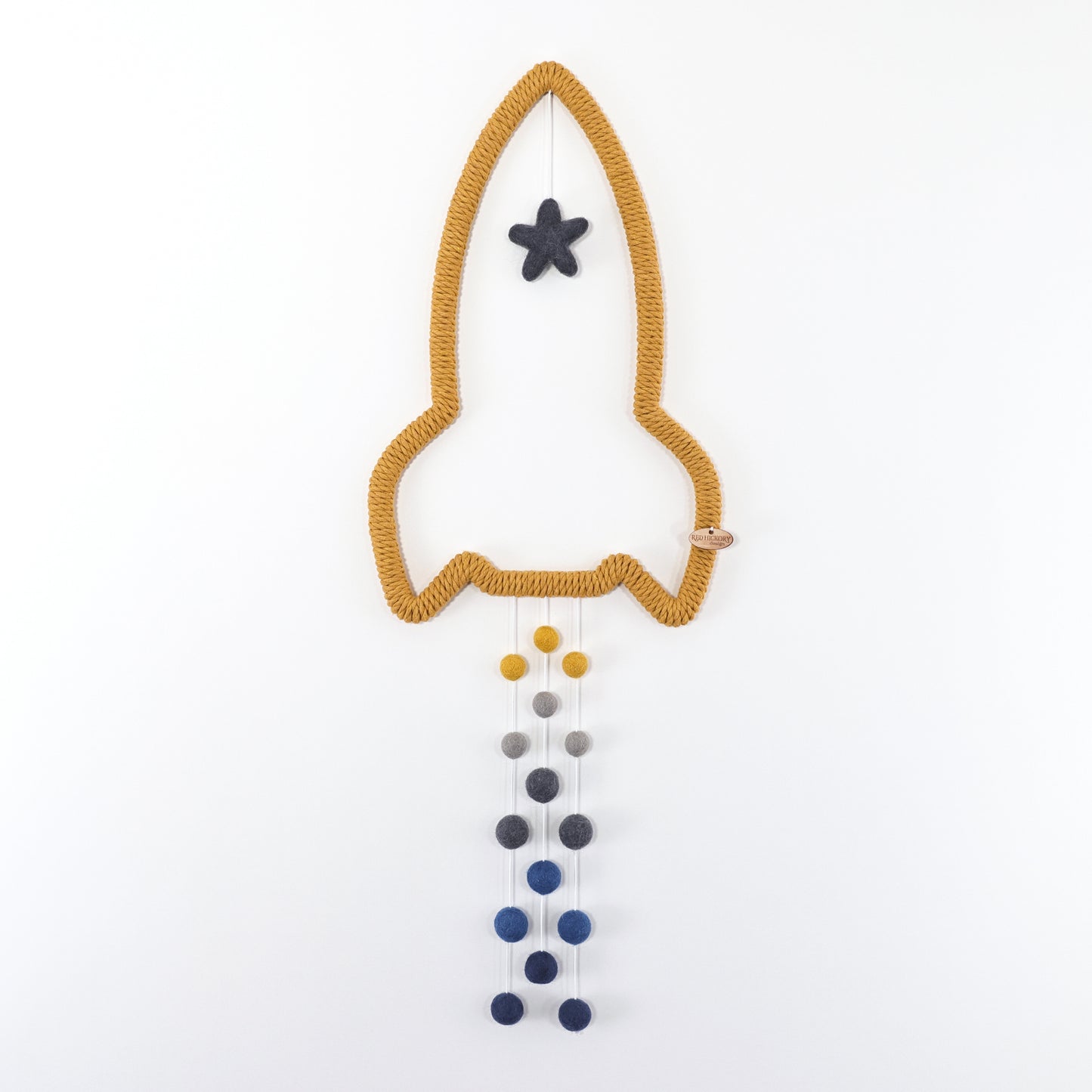 Yellow rocket shape wooden frame is wrapped with mustard yellow cotton yarn. Wool felt gray star hanging from the nozzle. Trails of wool felt pom-poms in blue, yellow, gray hanging from the bottom.