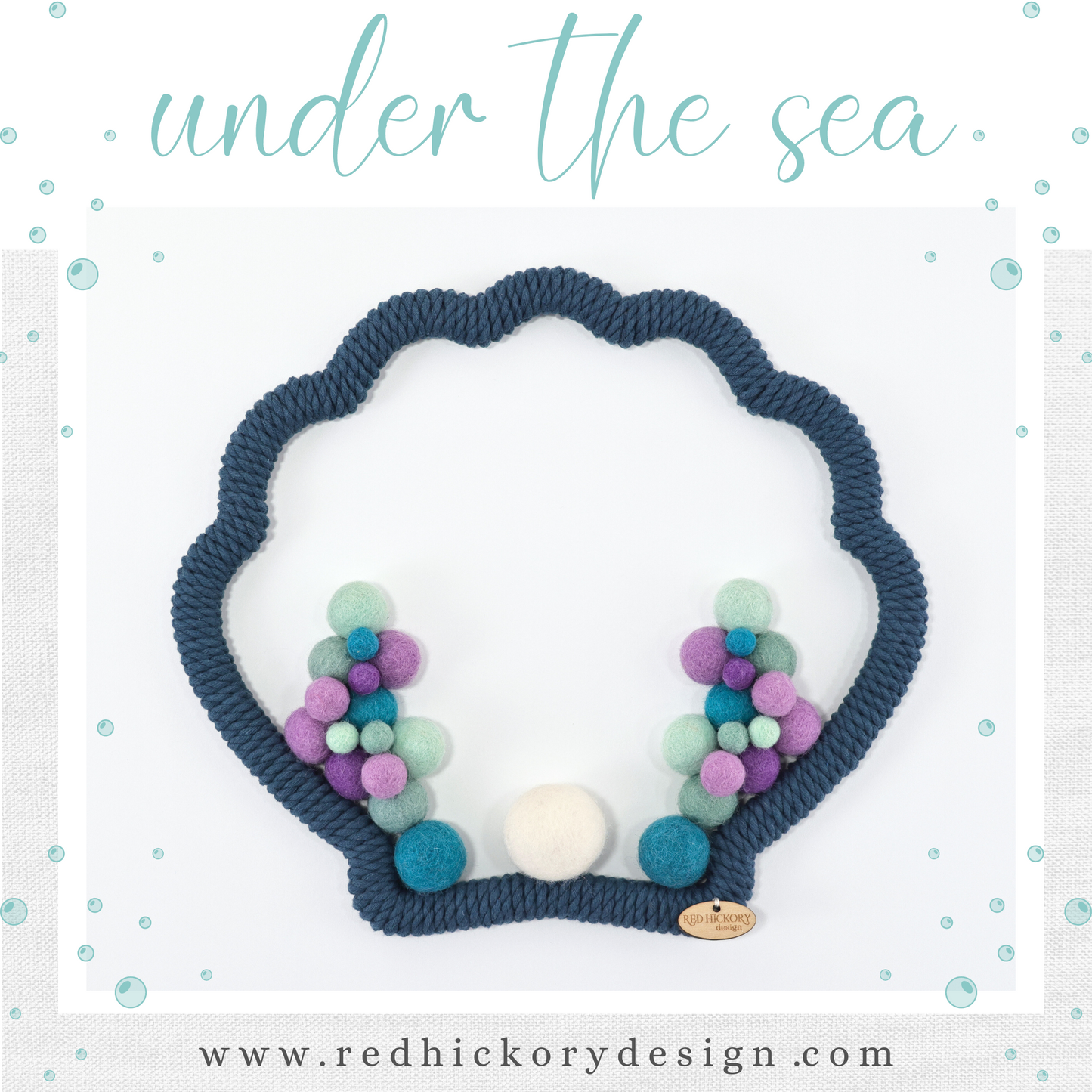 Sea shell shape wooden frame is wrapped with cotton teal color yarn. Soft wool felt pom-poms are attached to the inside of the frame. Colors are purple, light purple, teal, mint green and white. Wooden hand-stitched brand logo tag is hand sewn onto the bottom of the frame.